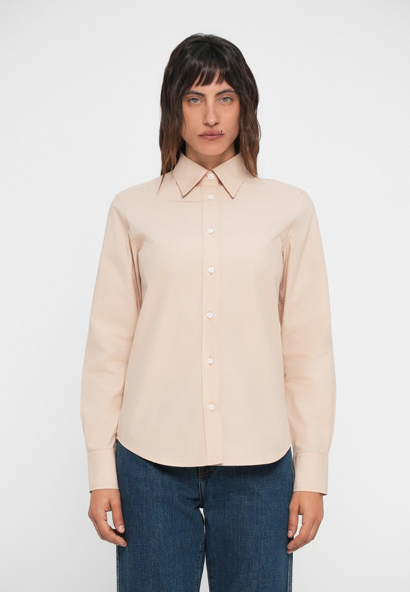 Beige button-up shirt with a pointed collar, long sleeves, and a smooth texture. Features a straight hem and is paired with dark jeans.