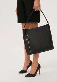 Fekete bőr kézitáska texturált felülettel, cipzáras zsebbel és arany tónusú fémalkatrészekkel. Egy kéz tartja, mellette egy fekete tűsarkú cipő látható.