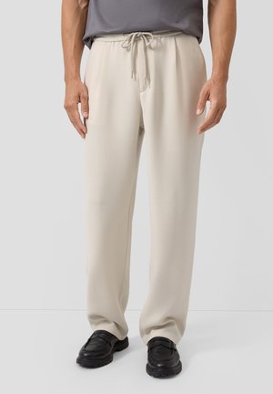 Beige broek met trekkoord gedragen met zwarte loafers en een ingestopt donkergrijs overhemd, zichtbaar van taille tot voeten.