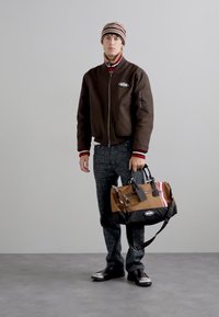 Brown bomber jacket with red and white accents, patterned pants, and a brown and black duffel bag with stripes and hardware details.