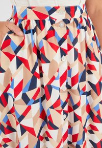 Jupe à motifs géométriques multicolores en tissu léger, présentant des teintes rouges, bleues, beiges et noires, avec des boutons sur le devant et des poches latérales.