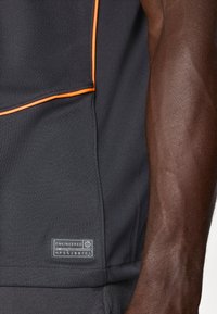 Svart sportsskjorte med oransje pipedetaljer. Har et teksturert stoff og en stoffetikett med ordet "ENGINEERED." Synlig atletisk arm.