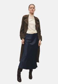 Manteau trench en suède marron porté sur un pull col roulé blanc, associé à une longue jupe en satin bleu marine et des bottes marron à hauteur de genoux. Le mannequin se tient les mains sur les hanches.