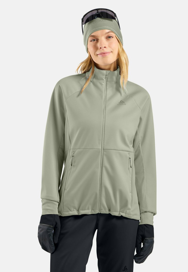 Frau trägt eine hellgrüne Zip-up-Jacke, eine passende Mütze mit Sonnenbrille obenauf, schwarze Handschuhe und schwarze Hose und steht vor einem weißen Hintergrund.