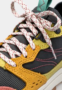 Colorful sneaker featuring grey mesh upper, yellow and orange suede accents, patterned white laces, and contrasting turquoise pull tab.