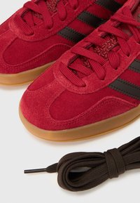 Piros velúr sportcipő fekete csíkokkal, lekerekített orral, texturált nyelvvel és barna gumitalppal. Egy barna cipőfűző van a cipők mellett.