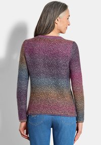 Femme aux longs cheveux gris portant un pull en tricot multicolore et un jean bleu, vue de dos.