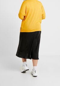 Top de punto amarillo con un corte relajado, combinado con una falda de satén negra y zapatillas blancas. Detalle de botones en el costado del top.