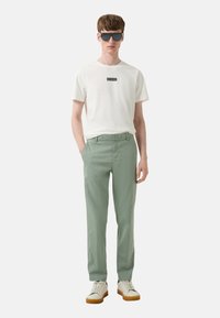 Witte t-shirt met zwart logo, lichtgroene broek en schoeisel met een witte bovenkant en een bruine zool; model draagt donkere zonnebril.