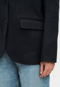 Κοντινό πλάνο ατόμου που φορά μαύρο μάλλινο σακάκι με τσέπη με καπάκι και λεπτομέρεια με κουμπί, σε συνδυασμό με ανοιχτό μπλε τζιν παντελόνι.