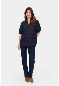 Marineblaue, leichte Bluse mit Lochstickerei, V-Ausschnitt und Puffärmeln. Kombiniert mit dunklen Denim-Jeans und beigen Ankle-Boots.