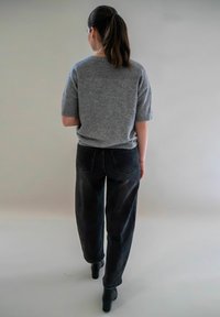 Femme avec des cheveux bruns attachés derrière, portant un pull gris à manches courtes, un jean noir et des bottines noires, s'éloignant sur un fond uni.