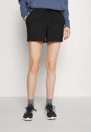 Osoba ubrana w czarne sportowe spodenki, niebieską bluzę, szare skarpety i czarne buty do biegania stoi na tle jednolitego jasnego tła.