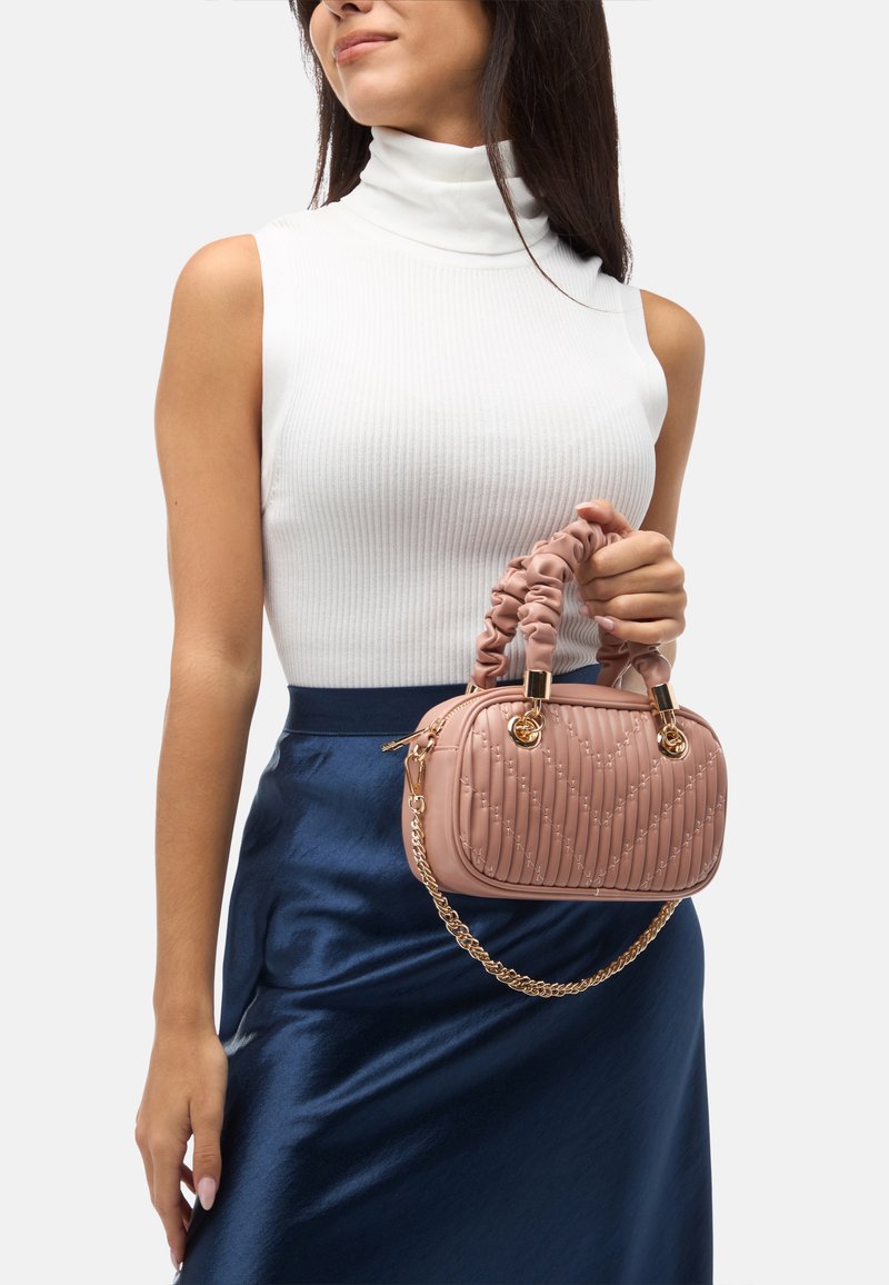 Rosa quiltad handväska med rund form, texturerad yta, skrumpna handtag och guldfärgade detaljer. Bärs med en vit topp och en blå kjol.