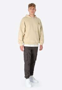 Beige übergroßer Hoodie mit einer Kängurutasche und gesticktem Logo, kombiniert mit dunklen Cargo-Hosen und weißen Sneakers.
