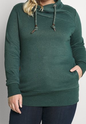 Osoba plus size nosící tmavě zelenou mikinu s kapucí se šňůrkami, dlouhými rukávy a jednou rukou v přední kapse, doplněnou tmavými džínami.