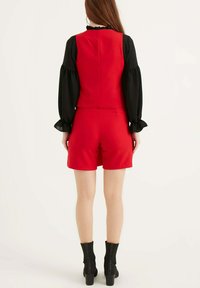 Romper rosso con busto aderente e pantaloni corti, abbinato a una maglietta nera a maniche lunghe con polsini a balze. Stivali neri alla caviglia completano il look.