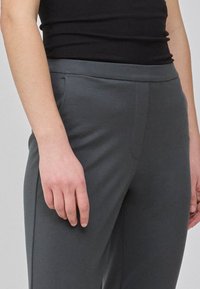 Grå elastiske bukser med en glat tekstur, front sømdetaljer og sidelommer, vist med en hånd hvilende på taljen.