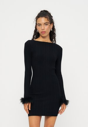 Femme aux cheveux bouclés portant une robe noire côtelée à manches longues avec des poignets en plumes, debout devant un fond clair uni.
