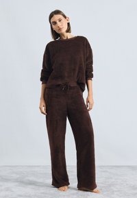 Braune, flauschige Lounge-Set aus weichem Stoff mit einem übergroßen Oberteil mit Umschlagsärmeln und Hose mit elastischem Bund. Die Textur ist plüschig und gemütlich.