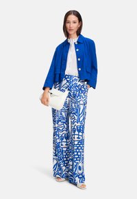 Blue linen jacket with gold buttons, white ruffled blouse, and high-waisted blue and white patterned wide-leg pants. Holding a white bag.