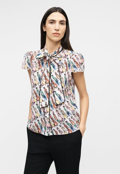 Kvinde med langt mørkt hår iført en farverig bluse med illustrationer og stort sløjfe, samt sorte bukser, stående med hænderne i lommerne.