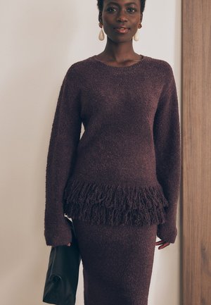 Burgundska pletena jopica z resicami na robu in dolgimi rokavi, v kombinaciji s usklajenim krilom. Tekstura je puhasta in mehka. Akcent črno torbico.