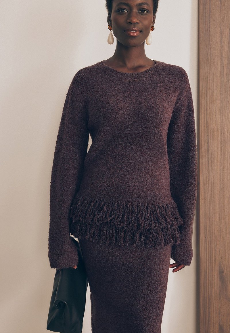 Burgundia lõngasviidiga sviiter, millel on fringi äär ja pikad varrukad, koos sobiva seelikuga. Tekstuur on kohev ja pehme. Must käekott täiendab.