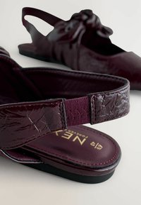 Burgundy slingback skor med en texturerad lackfinish. Funktioner inkluderar en slinga, elastiskt band och en slät innersula med guldtryck.
