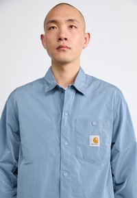Chemise à manches longues bleu clair en matière légère, avec une fermeture à boutons sur le devant, une poche poitrine et un écusson de logo visible.
