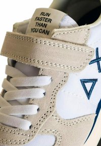 Scarpa sportiva bianca con accenti in suede beige, dotata di una fascia in Velcro, lacci bianchi e un'etichetta che recita "CORRI PIÙ VELOCE DI QUANTO PUOI."