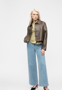 Young woman wearing a cropped brown leather jacket, olive green top, patterned wide-leg jeans, and black pointed heels, standing against a white background.