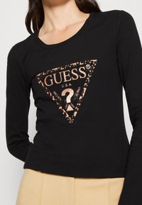 Schwarzes Langarmshirt mit einem dreieckigen Leopardenmuster-Logo in Roségold, versehen mit dem Markennamen "GUESS" und einem Fragezeichen-Detail.