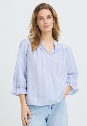 Žena v voľnej blúzke svetlomodrých pásikov s tromi štvrťovými nafúknutými rukávmi a svetlomodrých džínsach, stojaca pred jednoduchým pozadím.