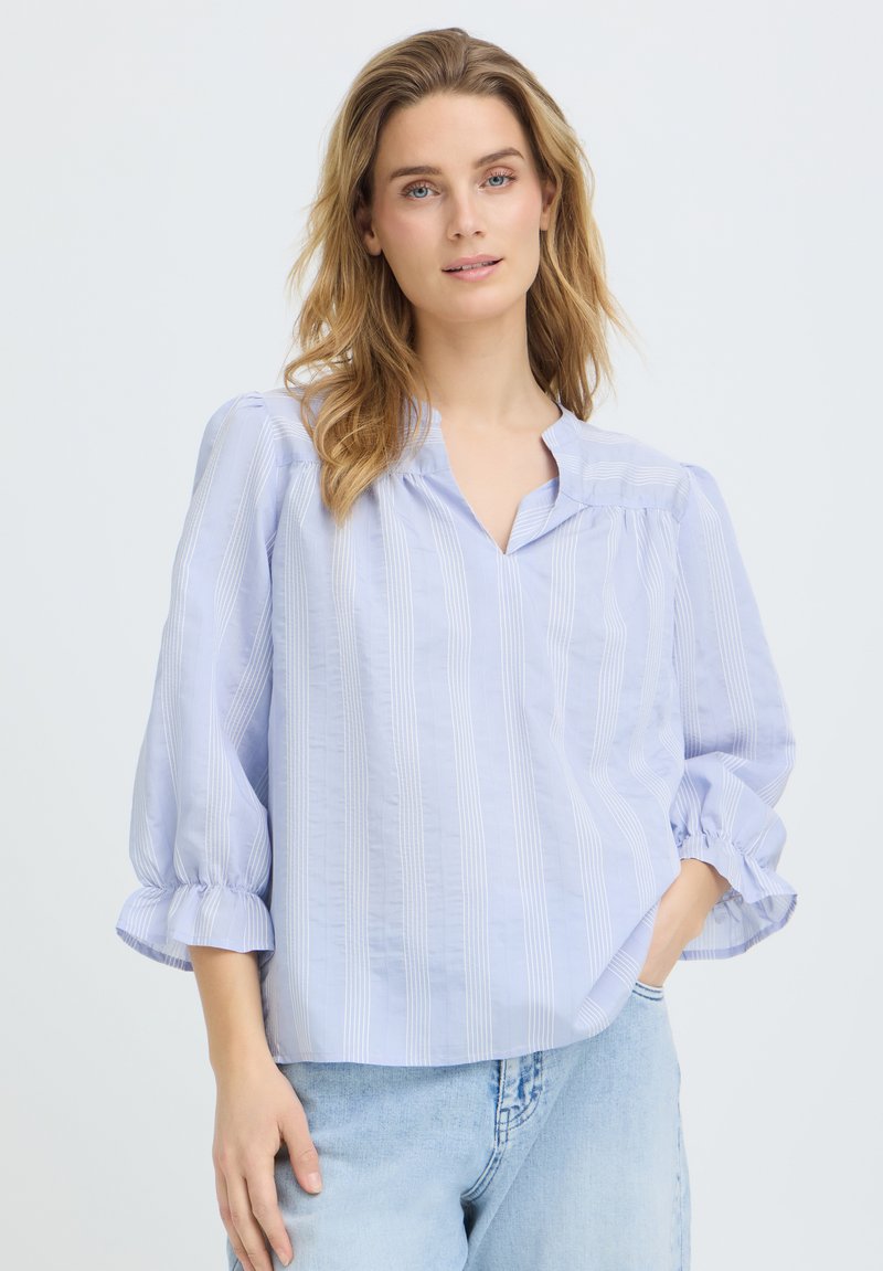 Nő laza, világoskék csíkos blúzzal, háromnegyedes puffadt ujjakkal és világoskék farmernadrágban, egyszínű háttér előtt állva.