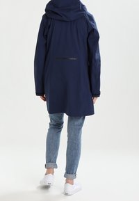 Navy waterproof jacket with a hood, featuring a rear zip pocket, and a straight cut. Paired with light blue cuffed jeans and white shoes.