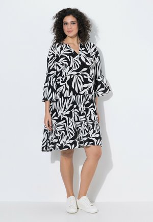 Kobieta o kręconych włosach, ubrana w czarno-białą sukienkę do kolan z liściastym wzorem i białe trampki, stojąca przy białej ścianie.