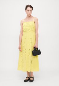 Un vestido maxi amarillo con bordados florales y recortes, tirantes finos y un corte relajado. Combinado con un bolso negro y zapatos negros con tachuelas.