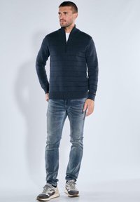 Marineblauer Pullover mit Reißverschluss am Kragen und horizontalen Streifen, kombiniert mit hellblauen Jeans und mehrfarbigen Sneakern. Glatte Textur und lässige Passform.