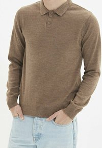 Polo marron à manches longues avec un ourlet côtelé, un col à trois boutons, et une texture douce, associé à un jean bleu clair.