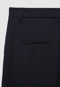 Primo piano di pantaloni blu navy che mostrano la vita, i passanti della cintura e una singola tasca a filetto sul retro su uno sfondo neutro.