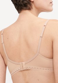 Beige podprsenka s nastaviteľnými ramienkami, hladkým materiálom a čipkovaným lemom pozdĺž pásu. Obsahuje zapínanie na chrbte pre úpravu prenikania.