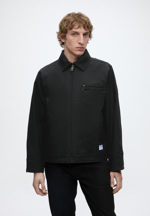 Mladý muž nosiaci čiernu bundu na zips s golierom a vreckom na hrudi na zips, stojaci pred jednoliatym bielym pozadím.