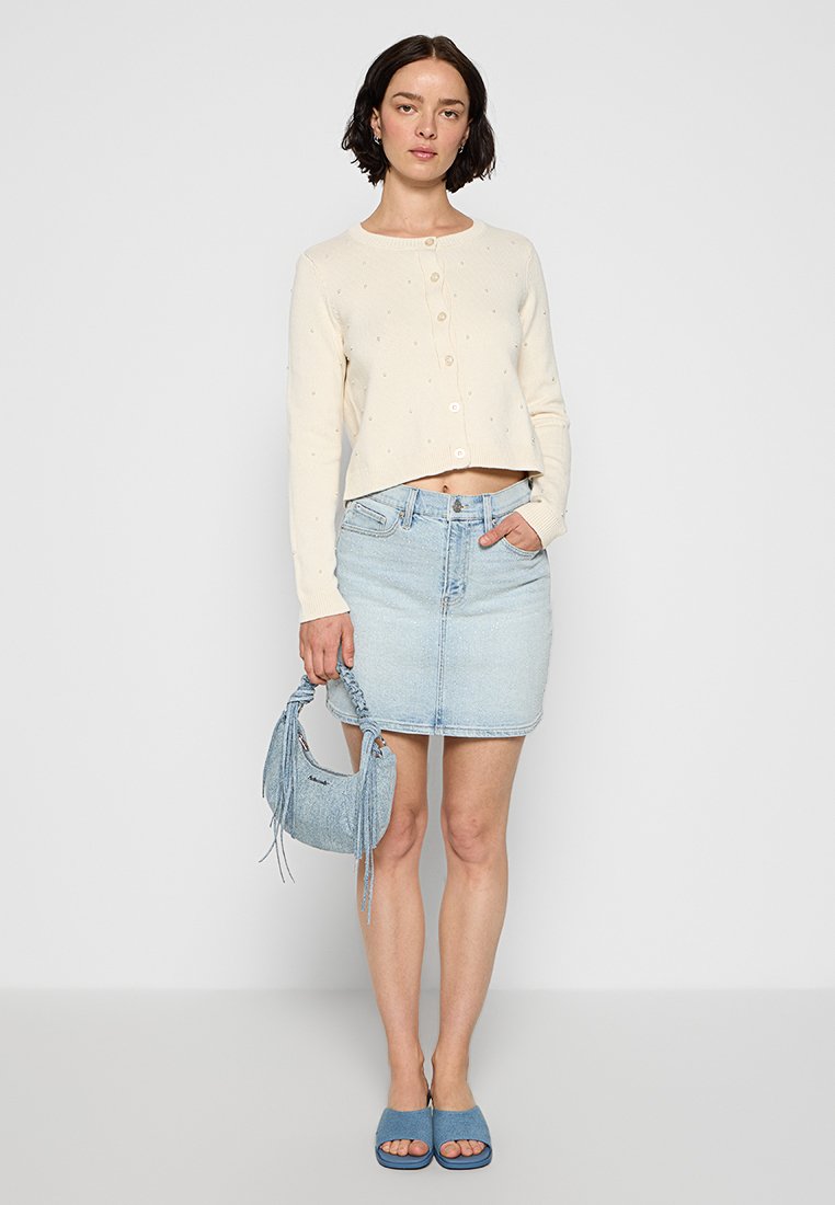 Hellbeige, gekürzter Cardigan mit Knöpfen und Struktur, kombiniert mit einem hellen Jeansrock, blauen klobigen Sandalen und einer fransigen blauen Handtasche.