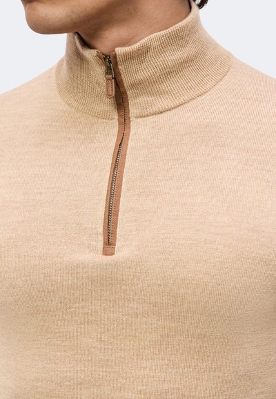 Pull en maille beige avec un col montant côtelé et une fermeture à glissière partielle à l'avant, porté par une personne montrée du cou vers le bas.