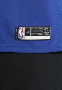 Blå baskettröja med nätstruktur, etiketten säger "NBA AUTHENTICS 44," inkluderar Nike-logotyp och texten "SWINGMAN." Svart nederkant.
