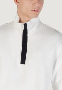 Sudadera blanca con cuello alto y media cremallera, que presenta un detalle de tela negra en el cuello. Textura suave y diseño moderno.