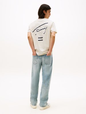 T-shirt de mangas curtas creme com impressão gráfica nas costas, combinado com jeans de corte solto azul claro e ténis brancos. Textura suave em todo o tecido.