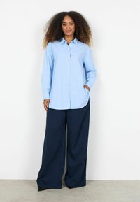 Sieviete ar cirtainiem matiem, ģērbusies gaiši zilā krekliņā ar pogām un platās tumši zilās biksēs, stāv pret balto, gludo sienu.