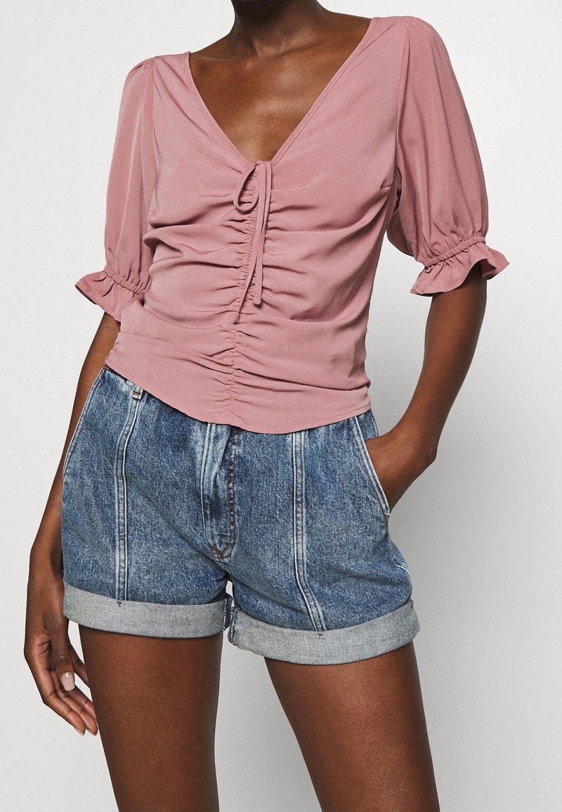 Blusa rosa empolvada con mangas cortas voluminosas y un frente fruncido, combinada con pantalones cortos de mezclilla de cintura alta con dobladillos enrollados.